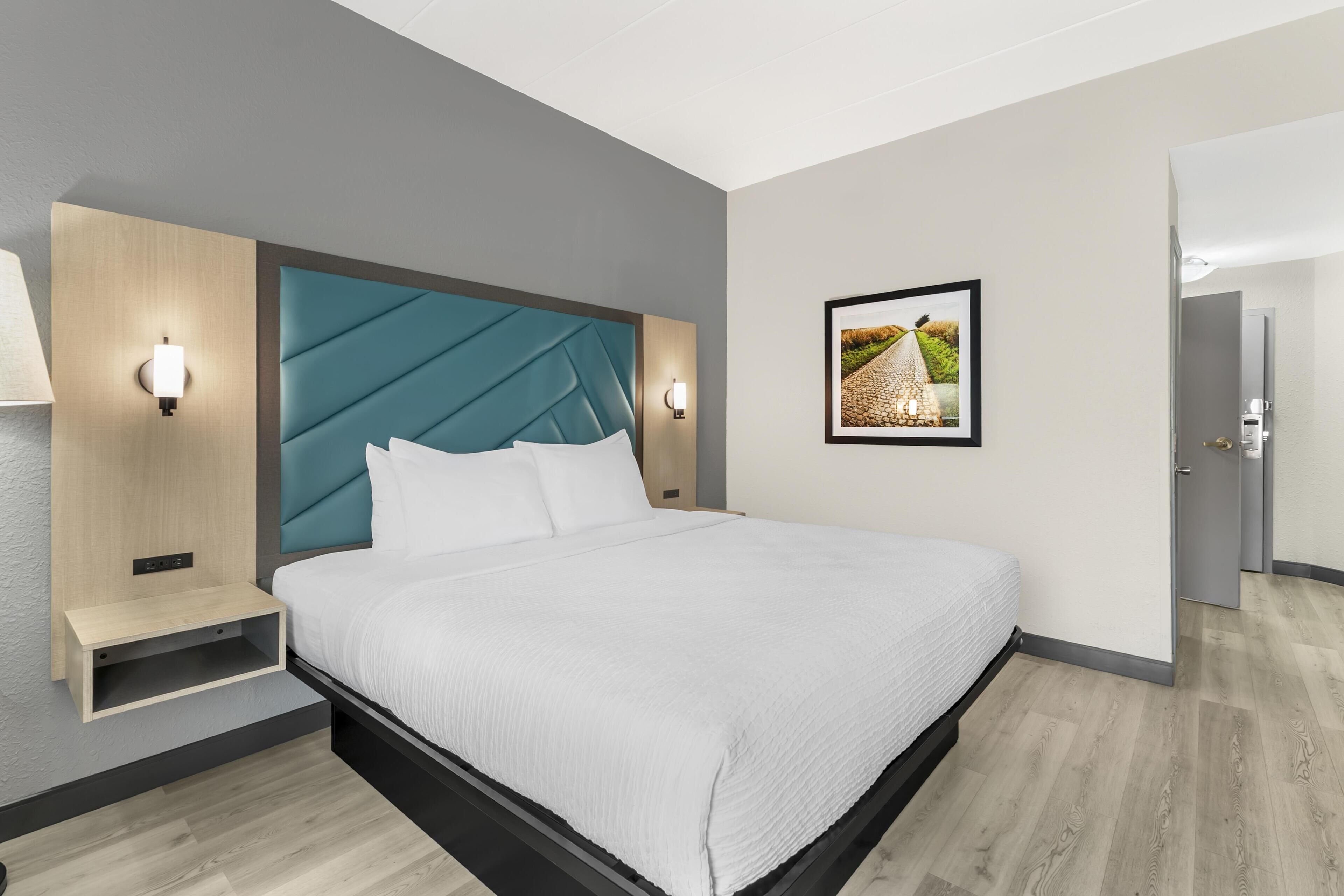 A modern hotel room with a bed, bedside tables, wall-mounted lights, a framed picture, and a door.