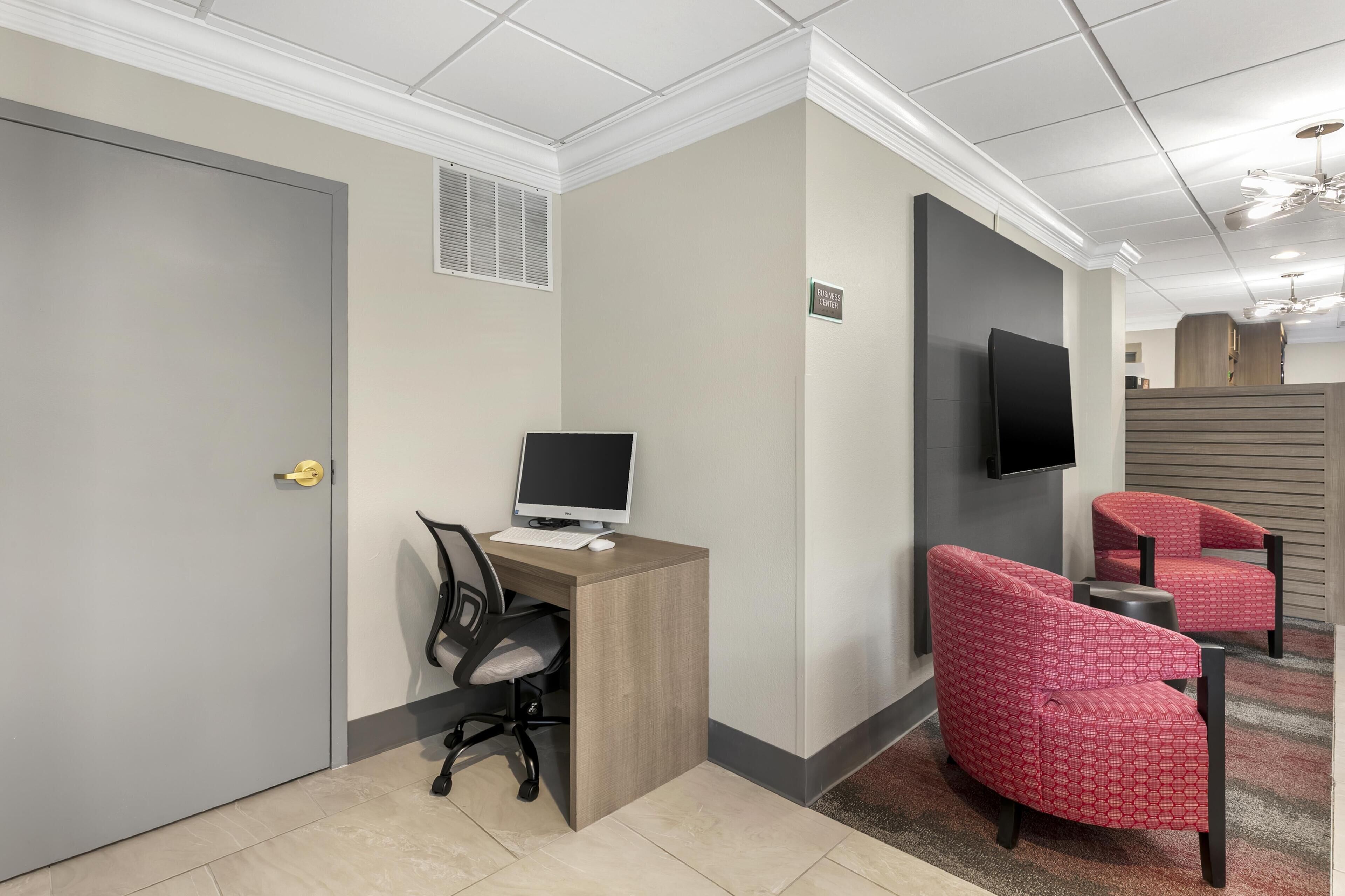 A hotel room with a desk, computer, two red armchairs, a television, and a door.