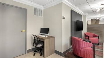 A hotel room with a desk, computer, two red armchairs, a television, and a door.