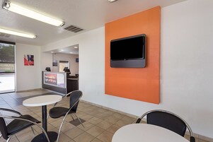 Um interior moderno com uma TV de tela plana fixada em uma parede laranja, um balcão de recepção e uma área de estar com mesas e cadeiras.