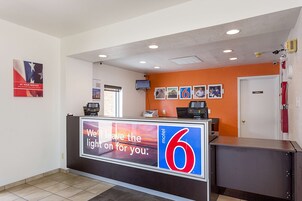 Uma área de recepção de hotel com uma grande placa com a mensagem "Deixamos a luz acesa para você" e o número "6" indicando o motel.