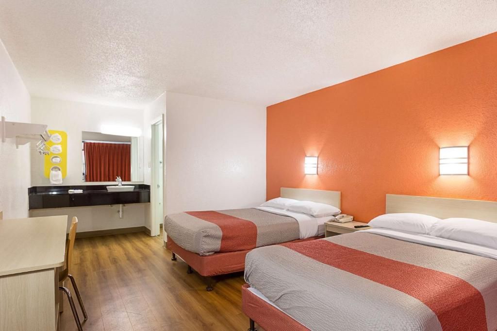 Een hotelkamer met twee bedden, een klein bureau, een spiegel en een badkamer met een rood gordijn.