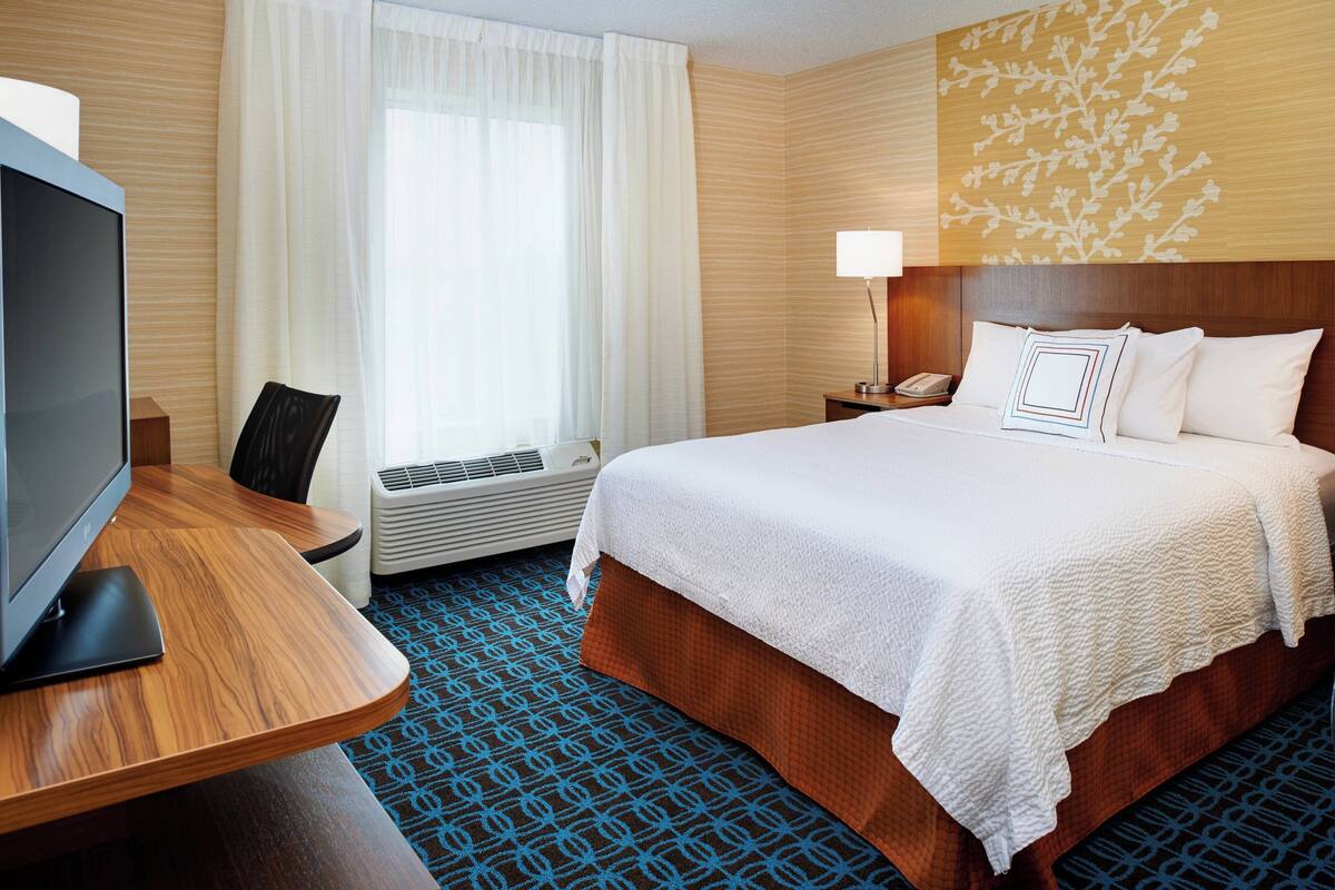A hotel room with a bed, a desk with a TV, a lamp, and a window with curtains.