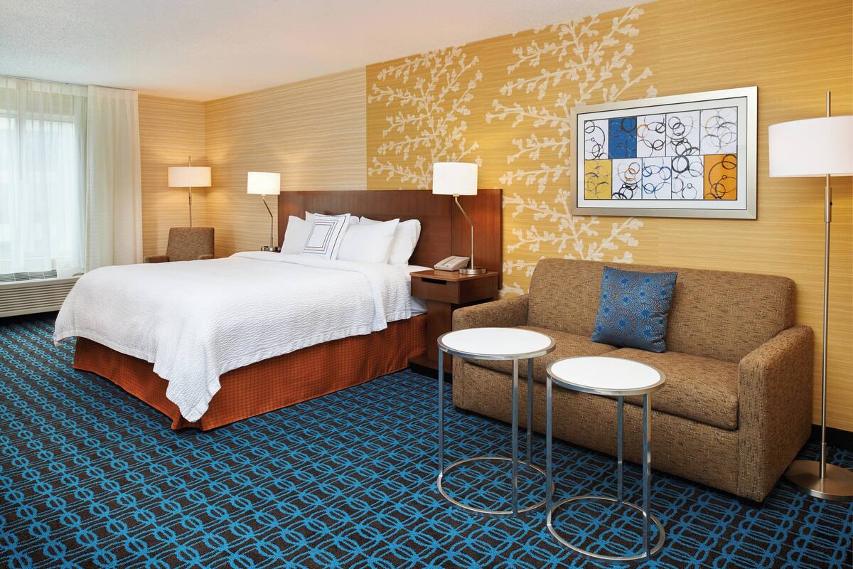 A hotel room with a large bed, a sofa, two small tables, and a wall art piece.