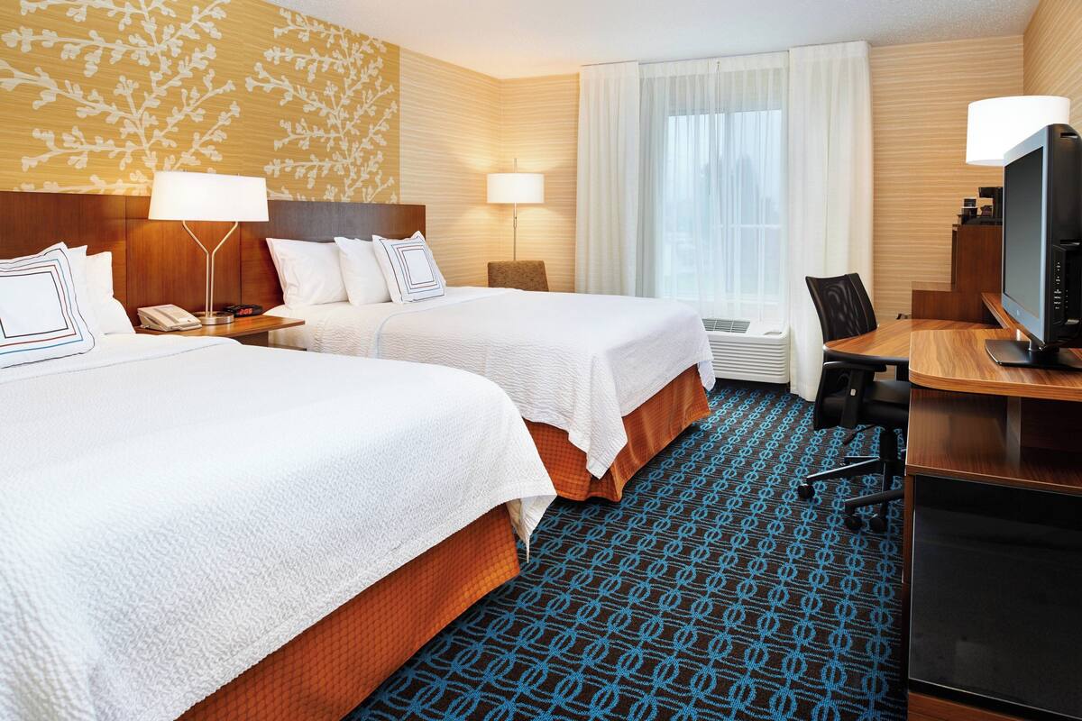 A hotel room with two beds, a desk, a TV, and a large window with curtains.