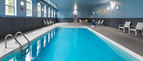 Una piscina con agua azul cristalina, rodeada de sillas y bancos blancos, y amplias ventanas a la izquierda.