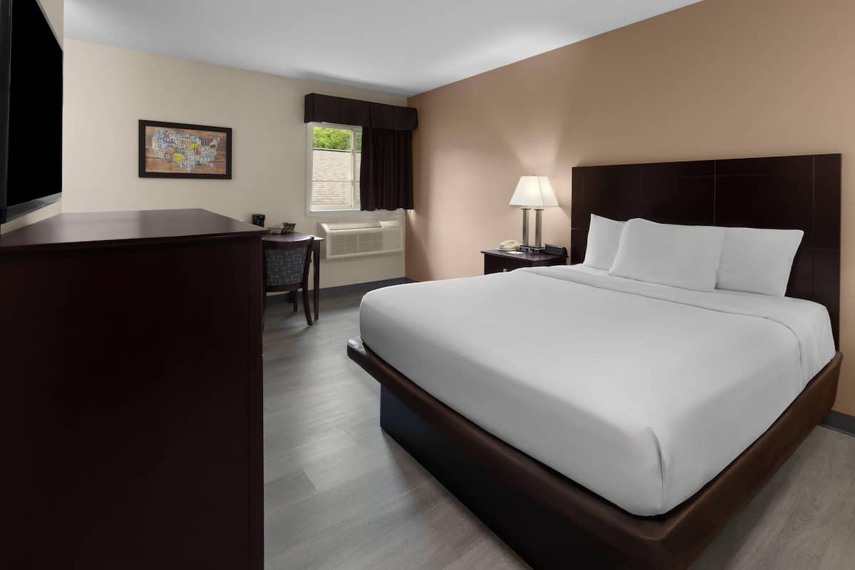 A hotel room with a large bed, a desk, a chair, a window with curtains, a wall-mounted TV, and a bedside lamp.
