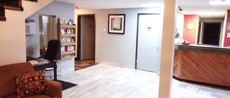 Um hall de hotel com uma mesa de recepção, um sofá com almofada estampada, uma estante de livros e uma porta que dá para outro quarto.