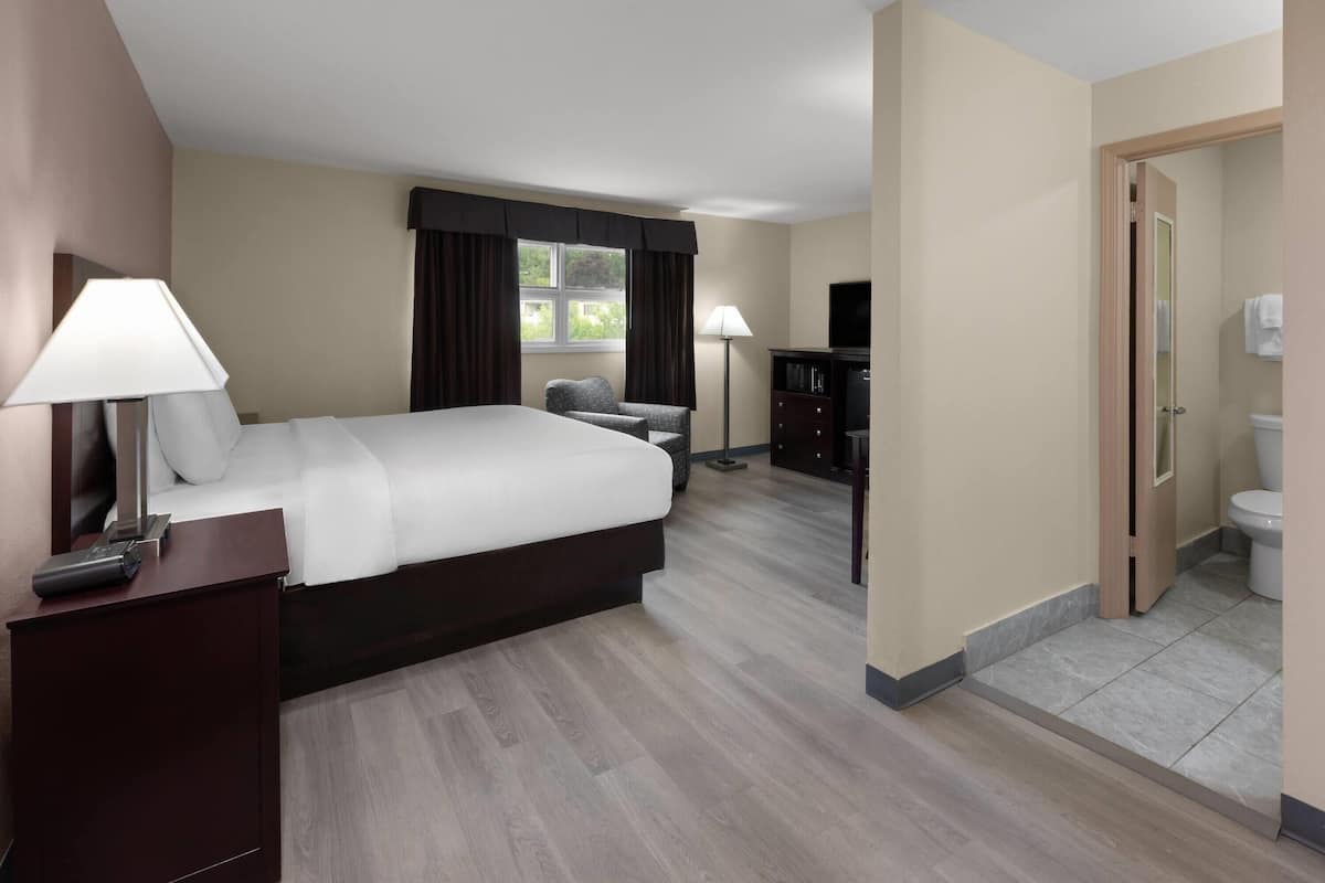 A hotel room with a bed, bedside table, lamp, chair, and a view of the outdoors through a window.