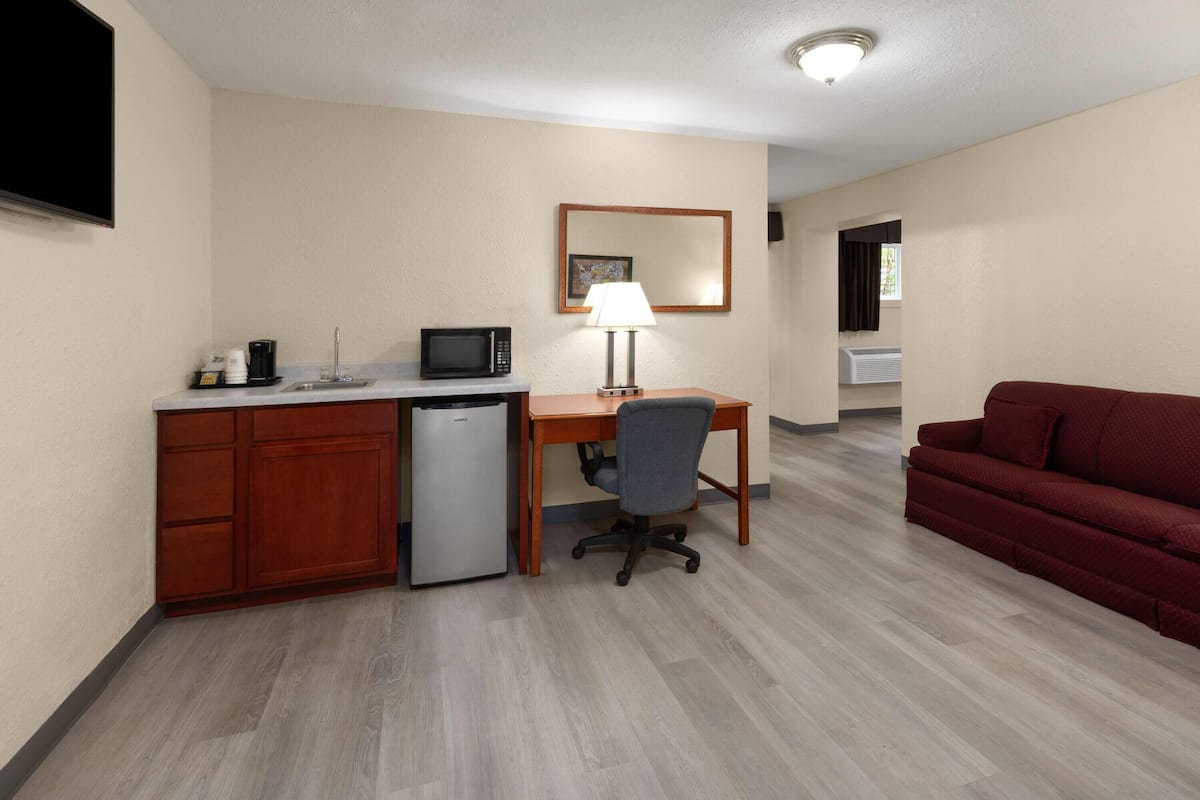 A compact hotel room with a kitchenette, a desk with a lamp, a red sofa, and a small refrigerator.