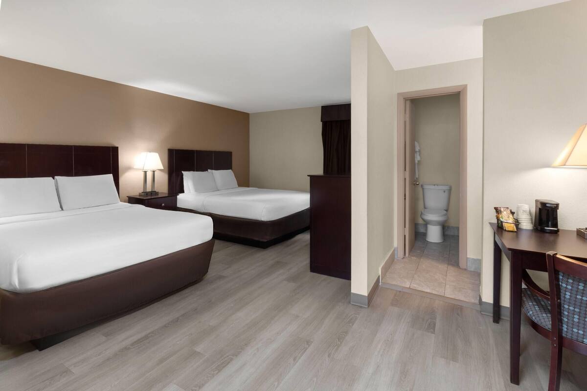 A hotel room with two beds, a desk, a chair, a lamp, and a bathroom visible through an open door.