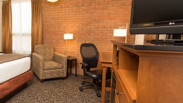 A hotel room with a bed, a chair, a desk, a television, and a brick wall.