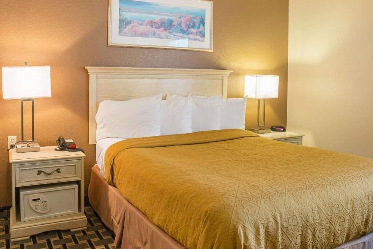 A hotel room with a bed, two bedside tables, a nightstand, a painting on the wall, and a lamp on each table.