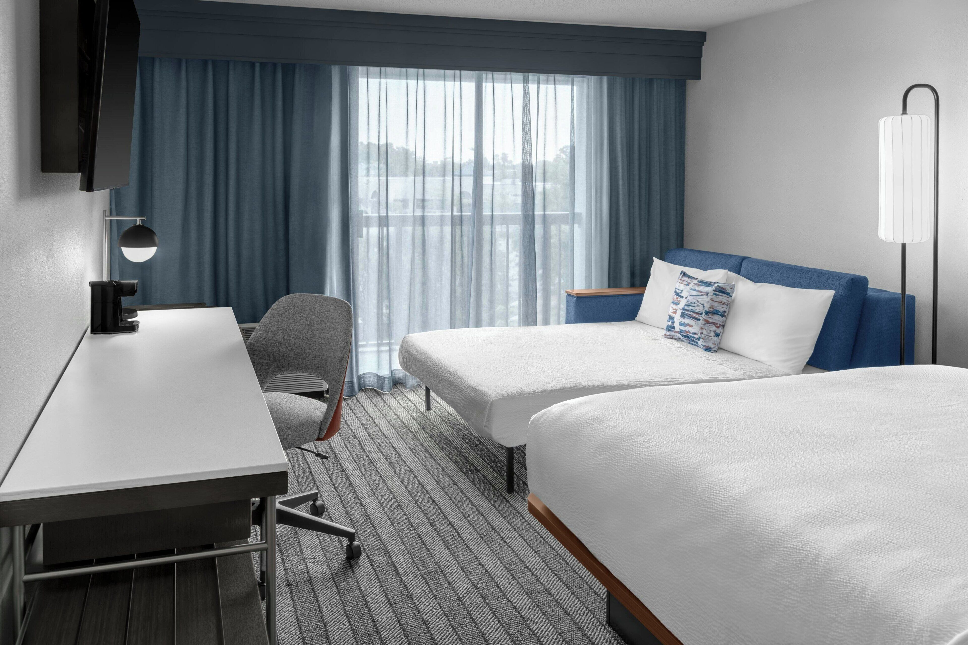 A hotel room with a desk, two beds, a chair, a lamp, and a view of the outdoors.