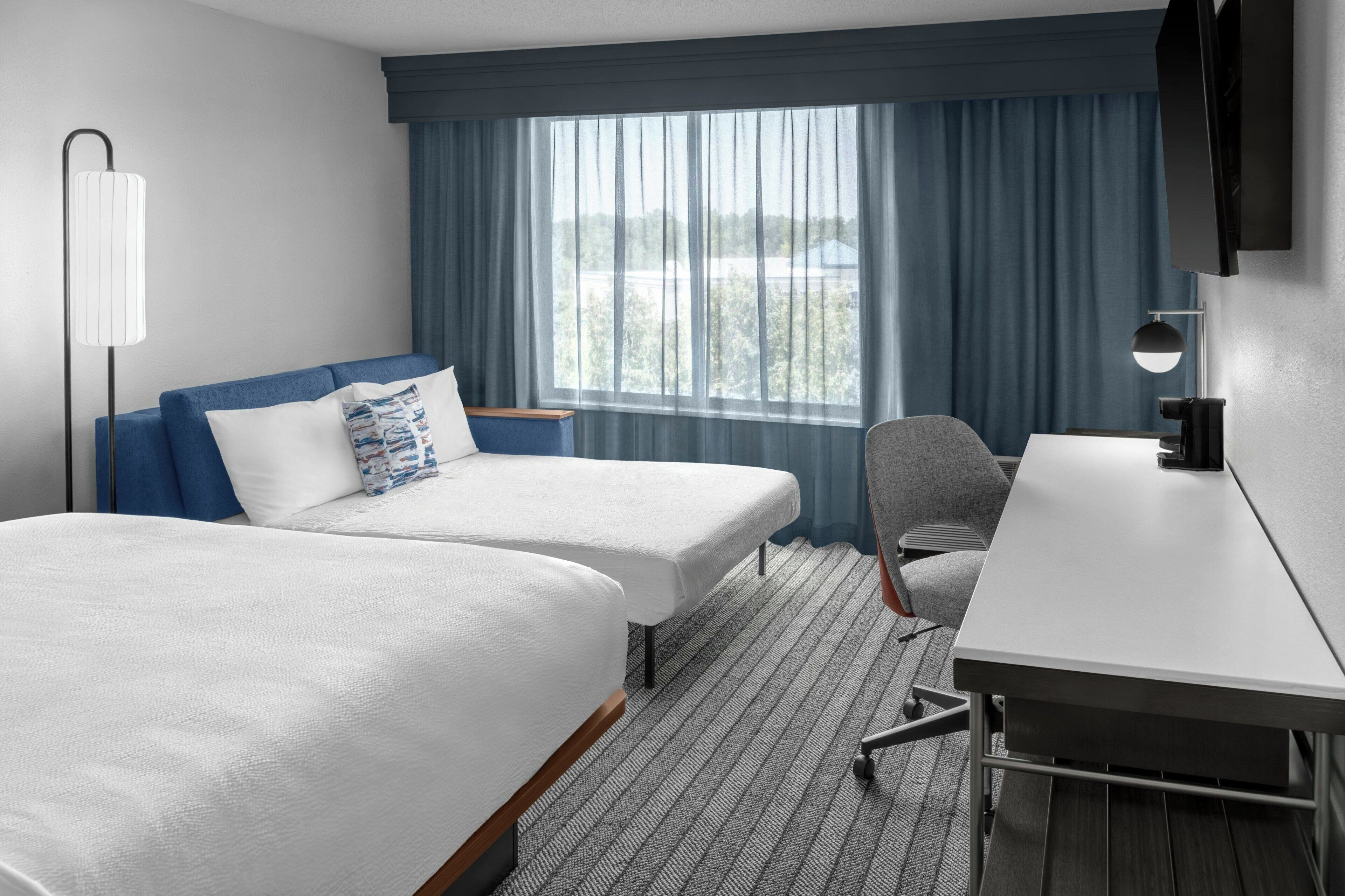 Une chambre d’hôtel avec deux lits, un bureau, une chaise, une télévision et une fenêtre avec des rideaux.
