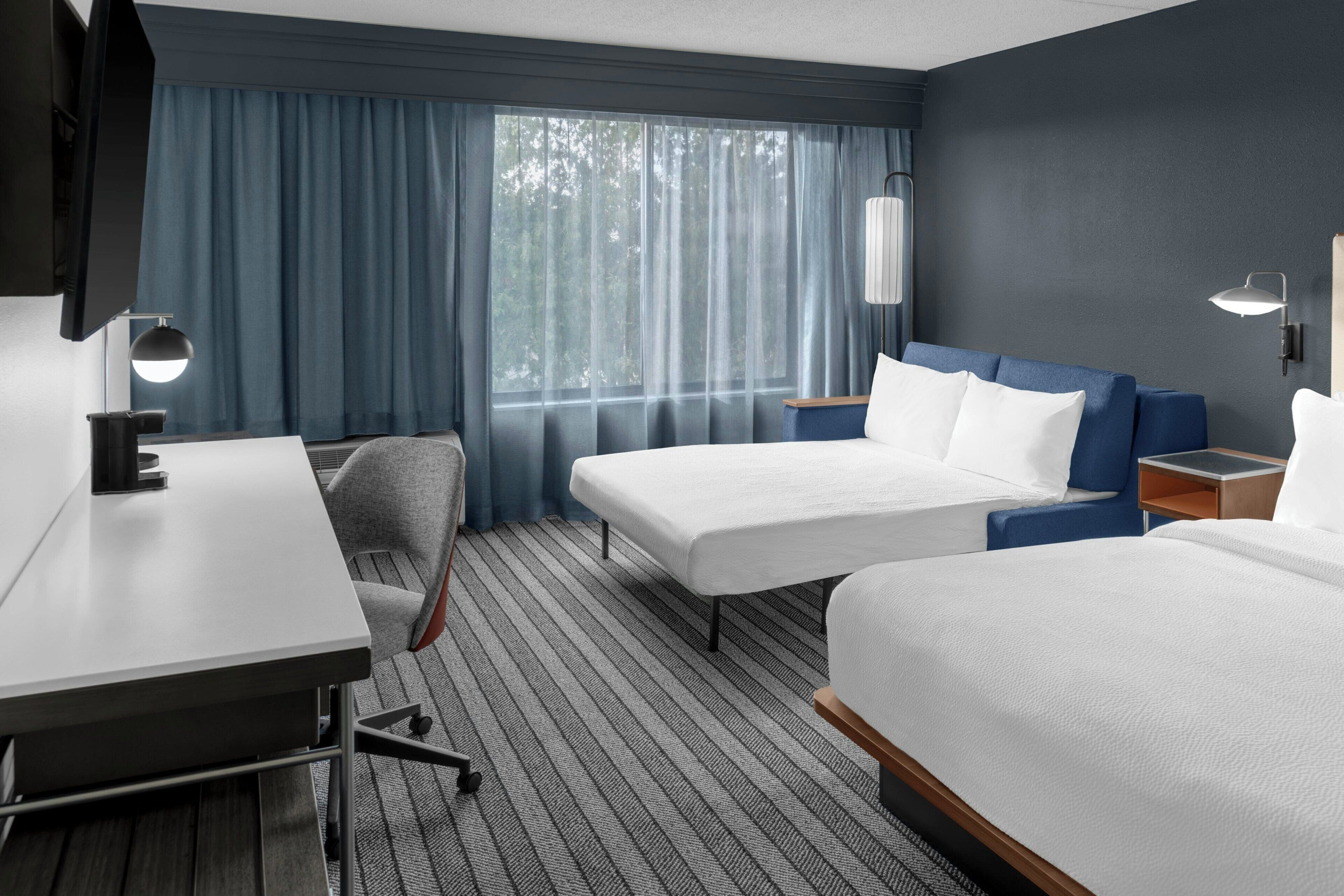 Habitación de hotel con escritorio, dos camas, una silla y ventana con cortinas.