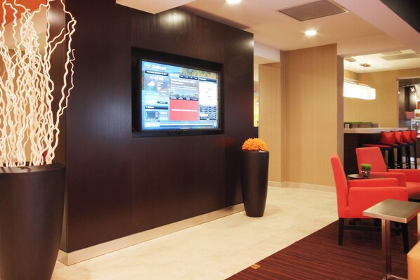 A modern lobby with a large flat-screen TV mounted on the wall, a reception desk, and a seating area with red chairs. - Courtyard Flint by Marriott (Flint)