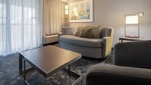Una habitación de hotel con un sofá gris, una mesa de centro de madera y una lámpara sobre una mesita auxiliar.