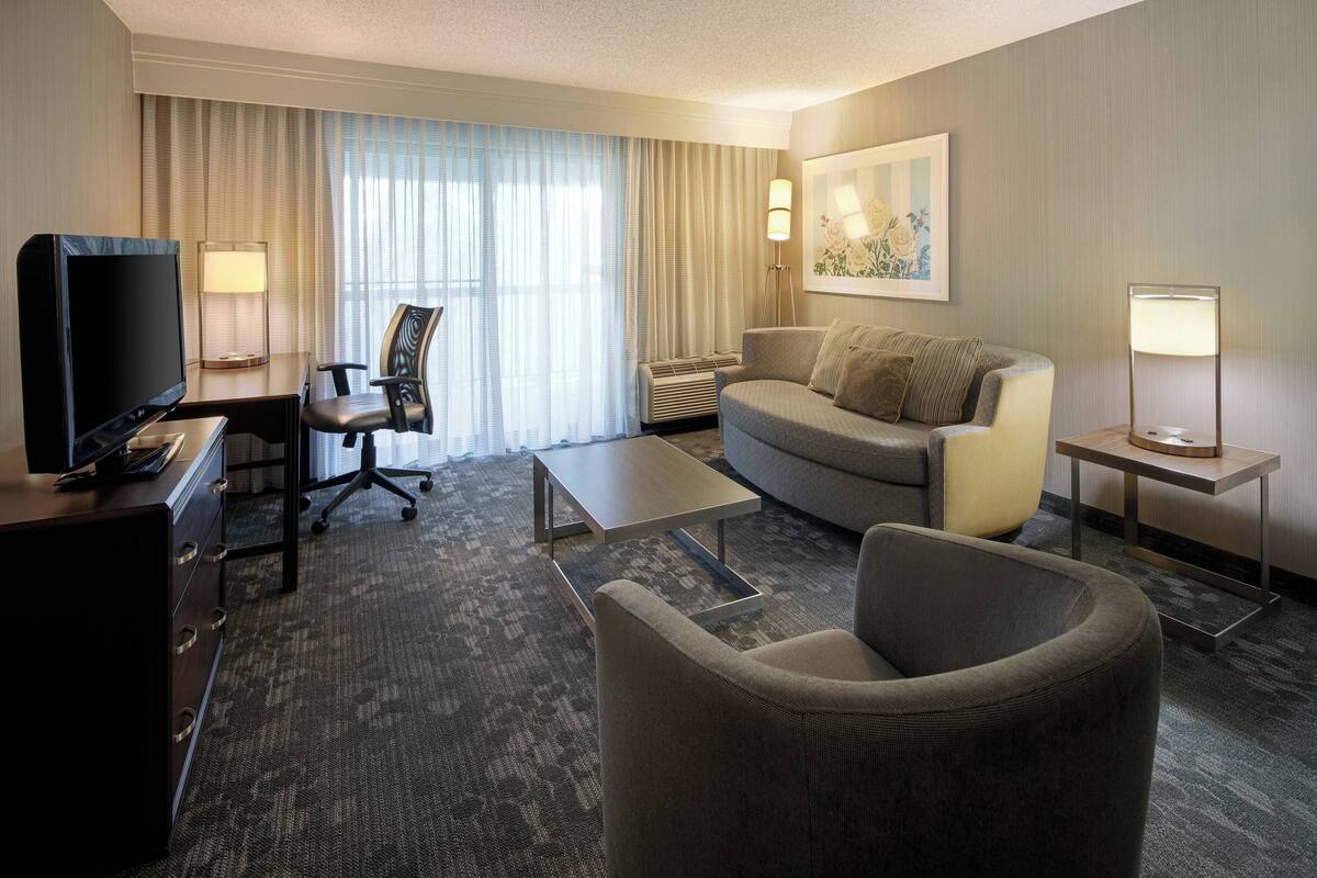 Habitación de hotel con televisión de pantalla plana, escritorio, silla, sofá, mesa de centro y dos lámparas de pie.