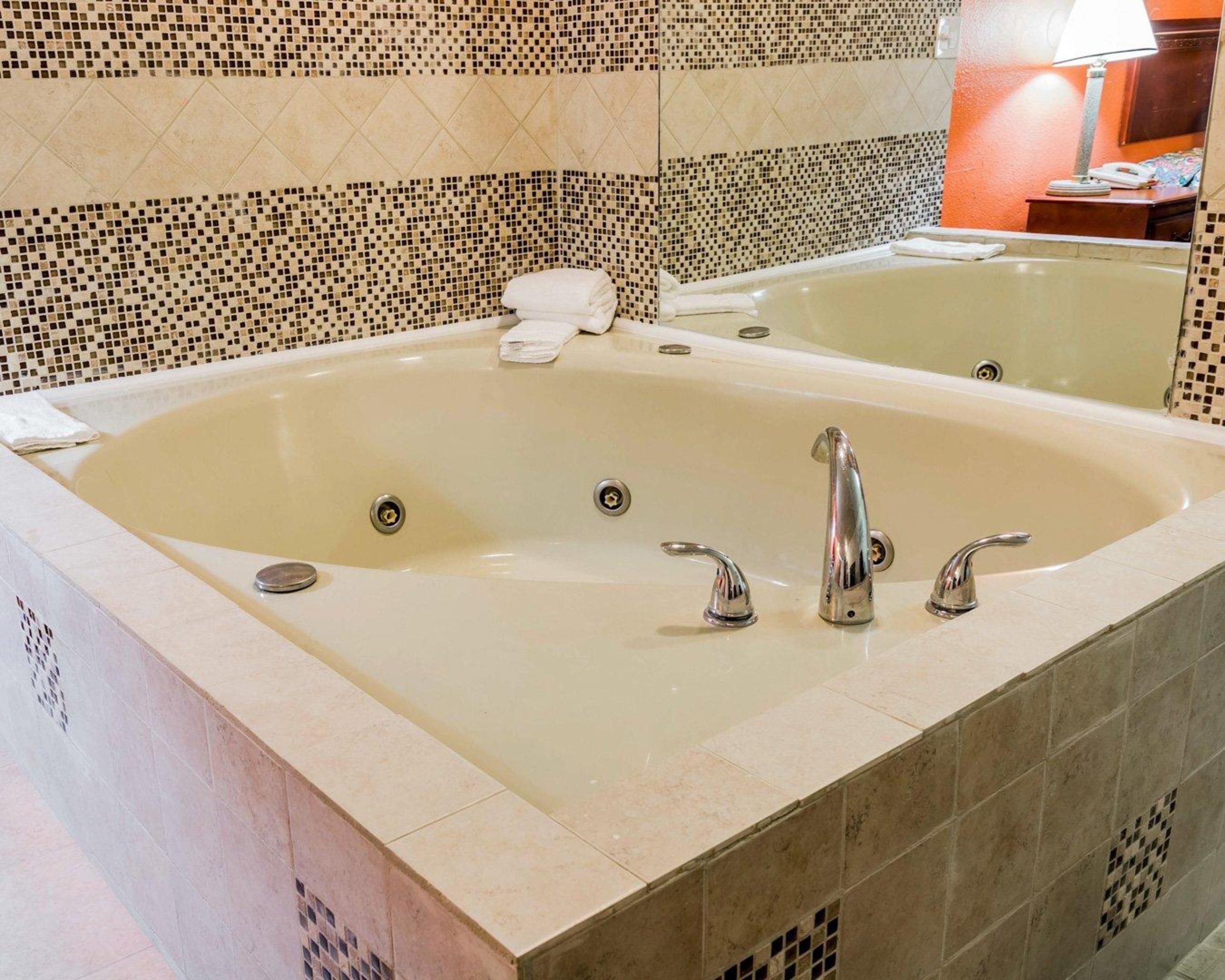 Une grande baignoire rectangulaire, dotée de deux robinets et d’un seul drain, entourée de carreaux de couleur beige avec une bordure en mosaïque.