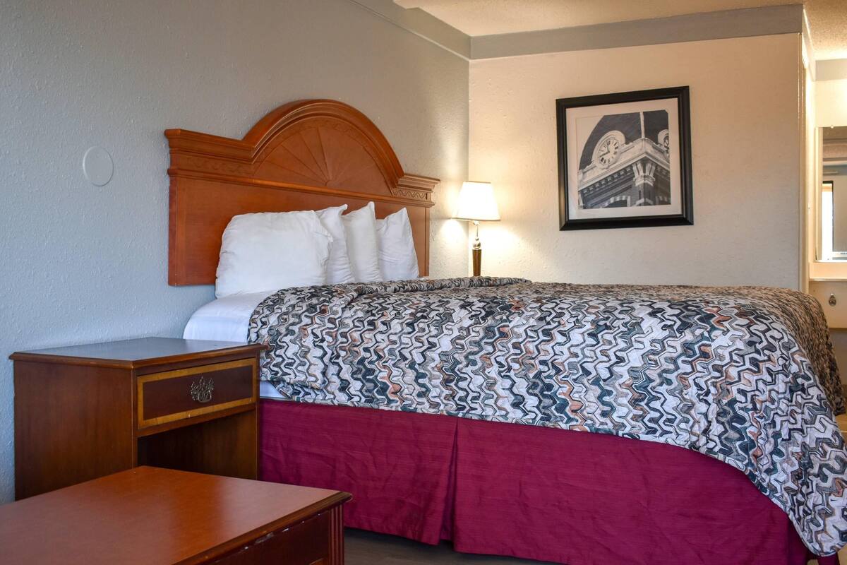A hotel room with a wooden headboard, a bed with patterned bedding, a nightstand, a lamp, and a framed picture on the wall.