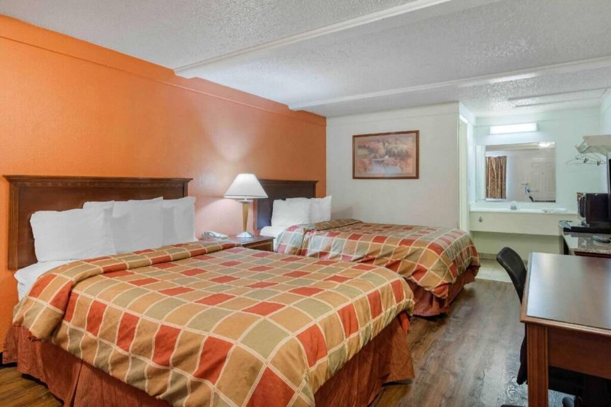 A hotel room with two beds, a desk, a chair, a lamp, and a painting on the wall.