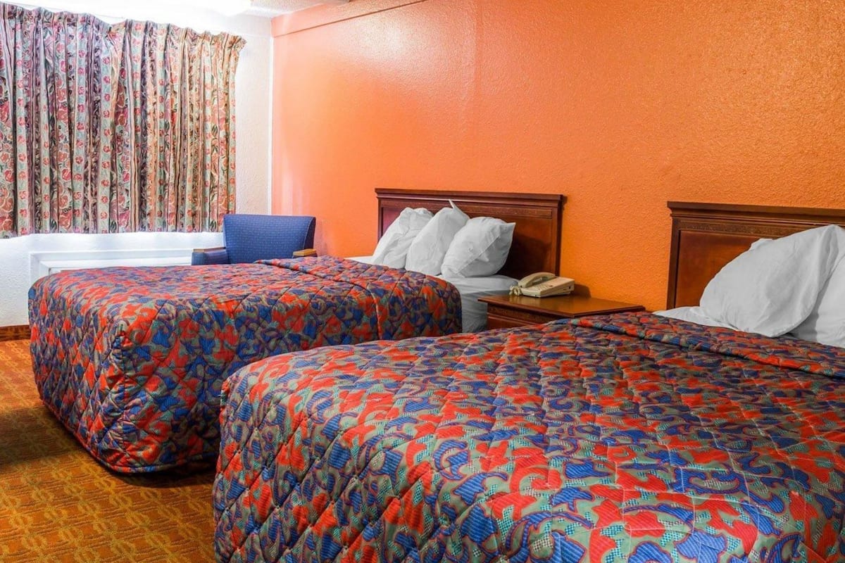 A hotel room with two beds, a blue chair, a wooden nightstand, and a window with curtains.