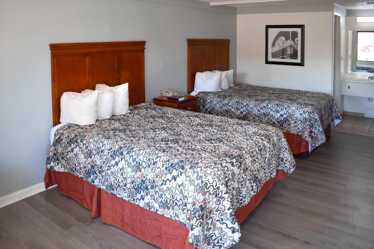 A hotel room with two beds, a wooden headboard, a bedside table with a lamp, a framed picture on the wall, and a window with a view of a building.