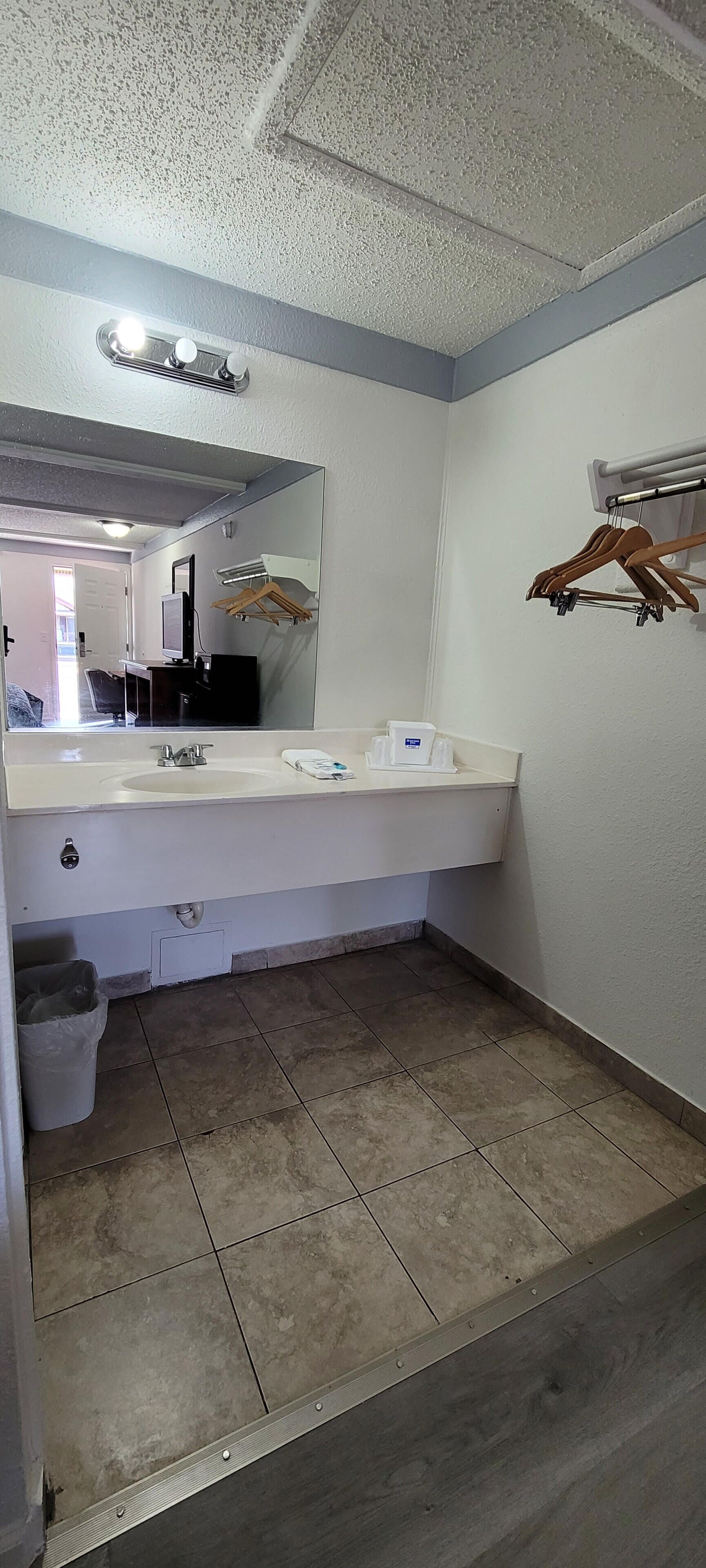 A bathroom with a white sink, mirror, and a towel dispenser. There is a trash bin and a wooden hanger rack on the wall.
