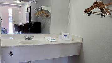 A bathroom with a white sink, mirror, and a towel dispenser. There is a trash bin and a wooden hanger rack on the wall.