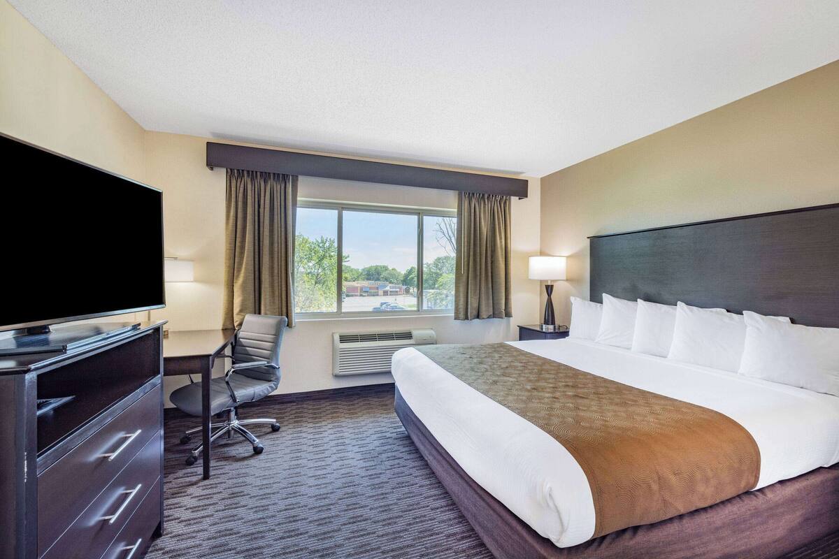 A hotel room with a large bed, a desk with a chair, a flat-screen TV, and a window with curtains.