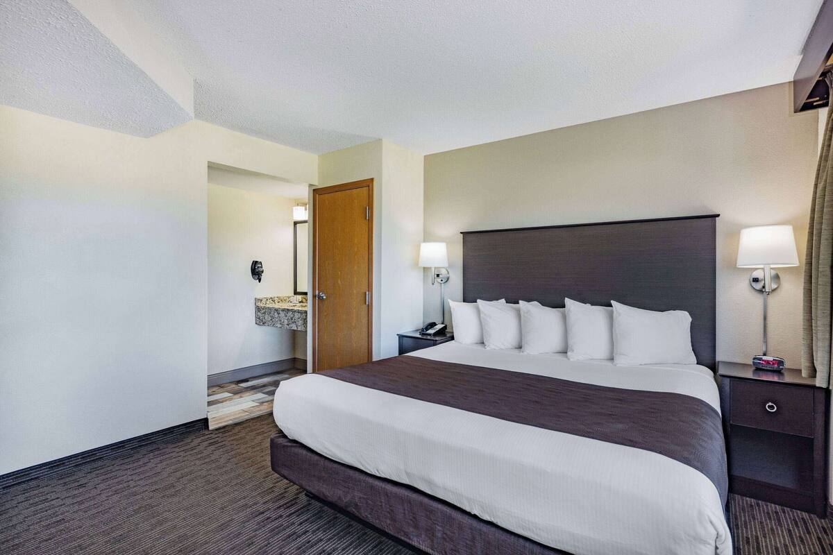 A hotel room with a large bed, a nightstand with a lamp, a wooden door, and a view of the hallway.