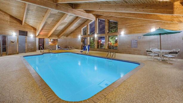 Une piscine intérieure avec des poutres en bois, des murs carrelés et un espace détente équipé d’un parasol.