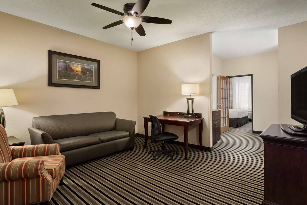 A hotel room with a sofa, armchair, desk, lamp, television, and a ceiling fan.