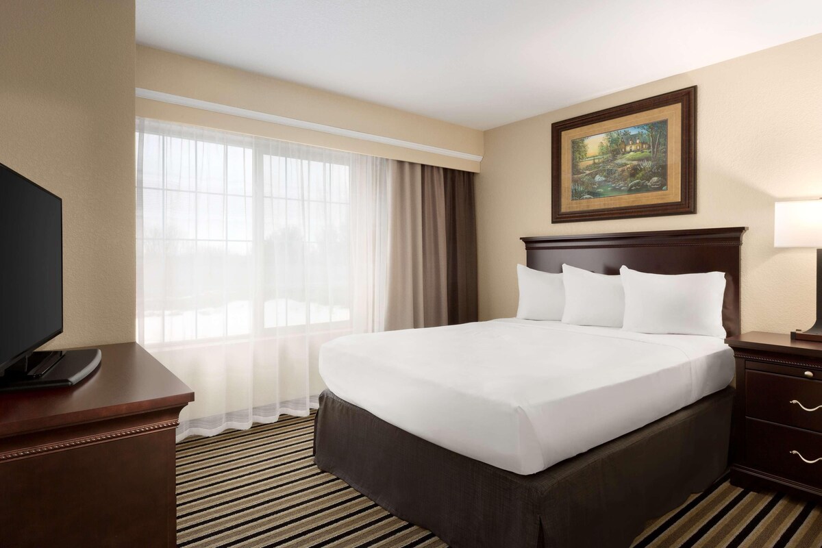 A hotel room with a large bed, a television on a stand, a painting on the wall, and a window with curtains.