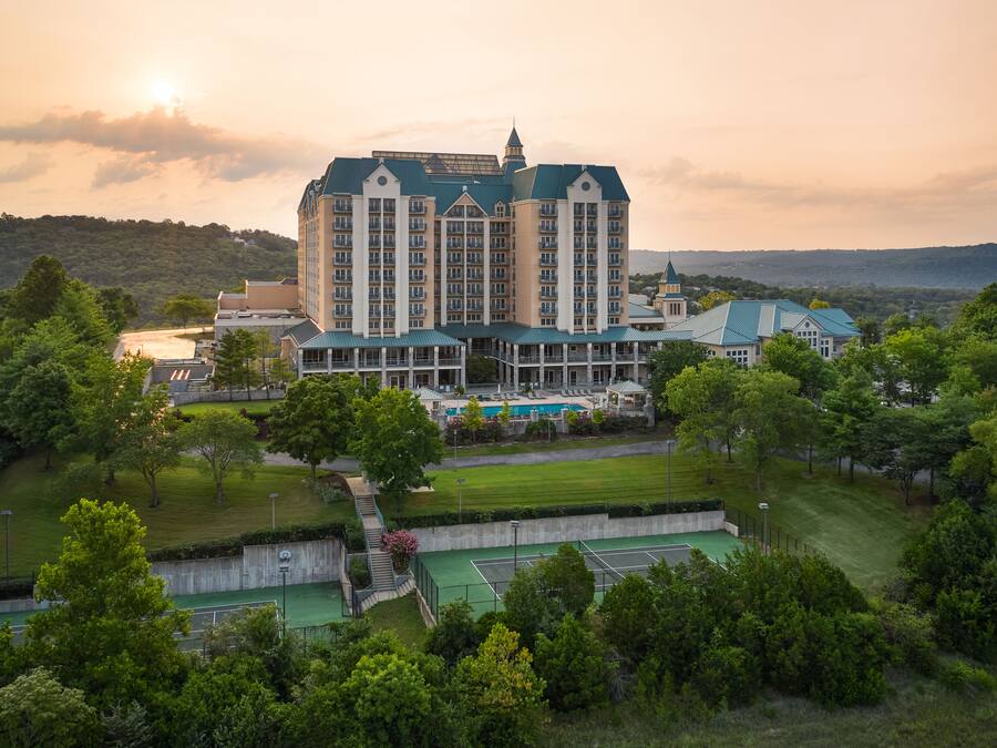 A large hotel with multiple stories, a swimming pool, tennis courts, and lush greenery surrounding the property.