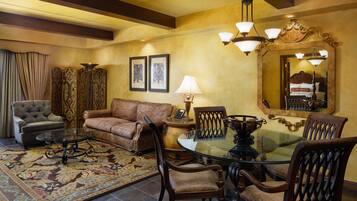 A dining area with a glass table, wooden chairs, a patterned rug, a sofa, a lamp, and framed artwork on the wall.