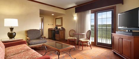 A hotel room with a sofa, armchair, coffee table, television, and a view through the door.