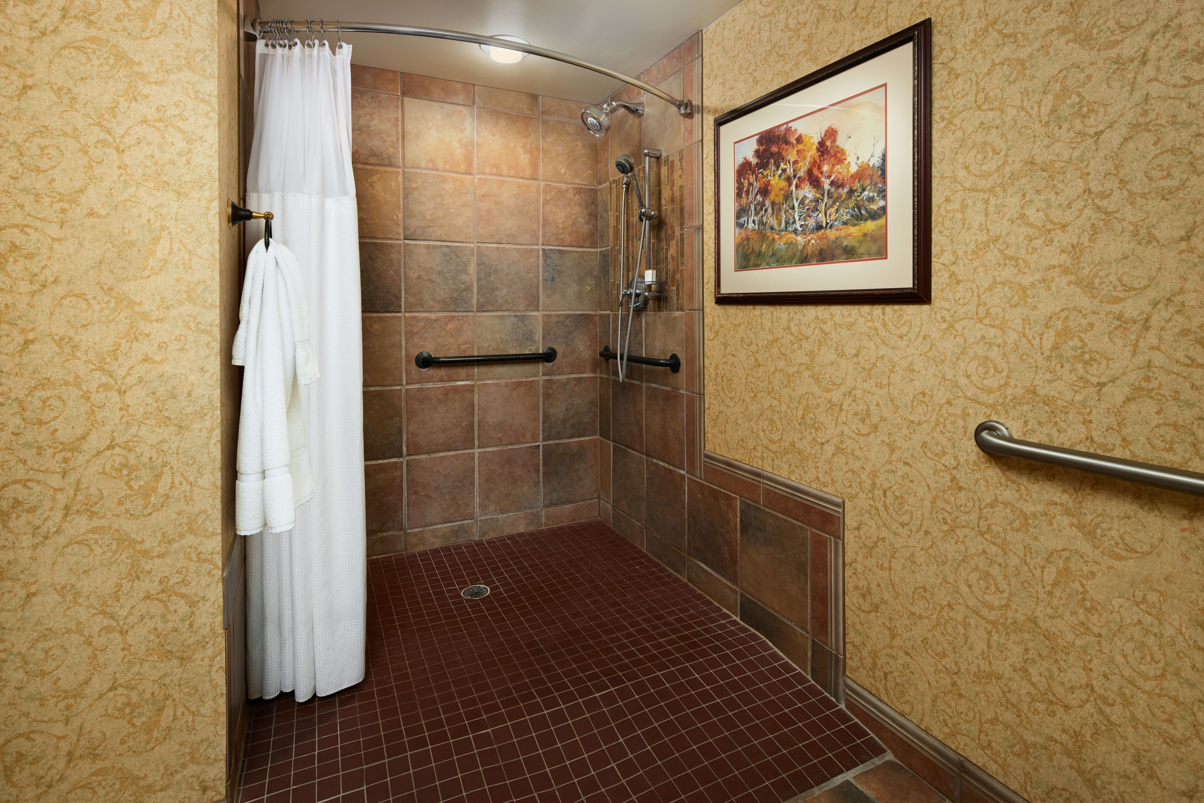 A shower area with a tiled floor, a white shower curtain, a framed picture on the wall, and a handrail.