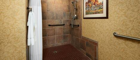 A shower area with a tiled floor, a white shower curtain, a framed picture on the wall, and a handrail.