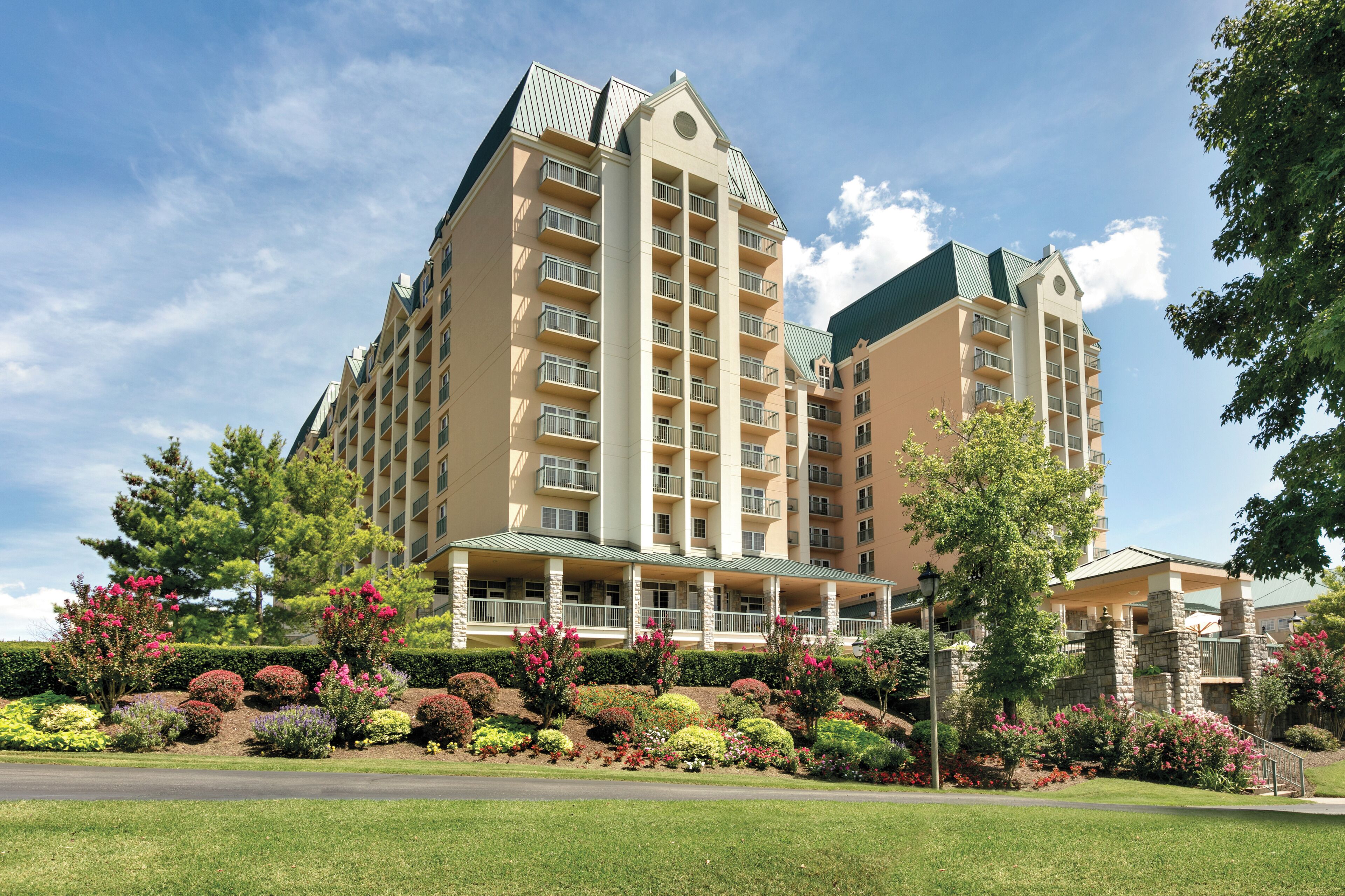 A multi-story hotel with balconies, a covered entrance, and landscaped gardens with various shrubs and flowers.