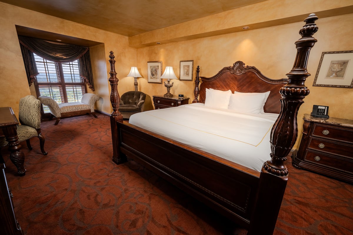 A spacious bedroom with a large bed, a sitting chair, a desk, and a window with curtains.