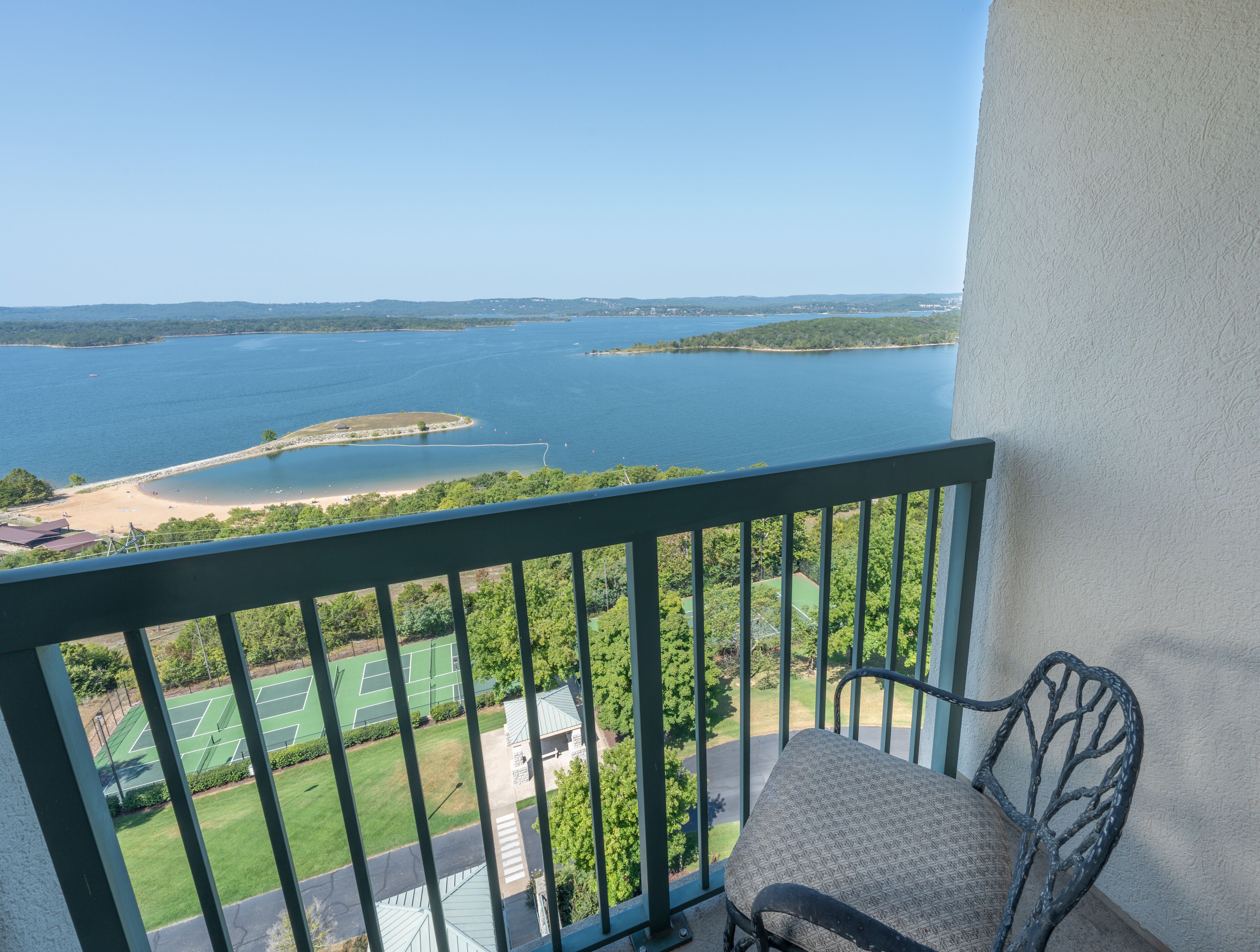 A balcony with a view of a lake, a tennis court, and a chair.