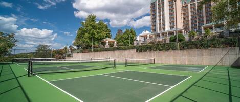 Una cancha de tenis con red, rodeada de un muro de piedra y vegetación, con un edificio de hotel al fondo.