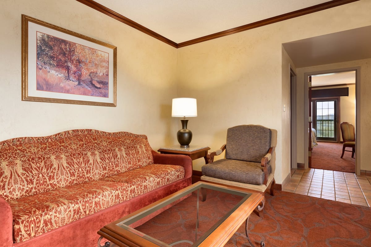 A living room with a patterned sofa, a chair, a glass coffee table, a lamp, and a framed painting on the wall.