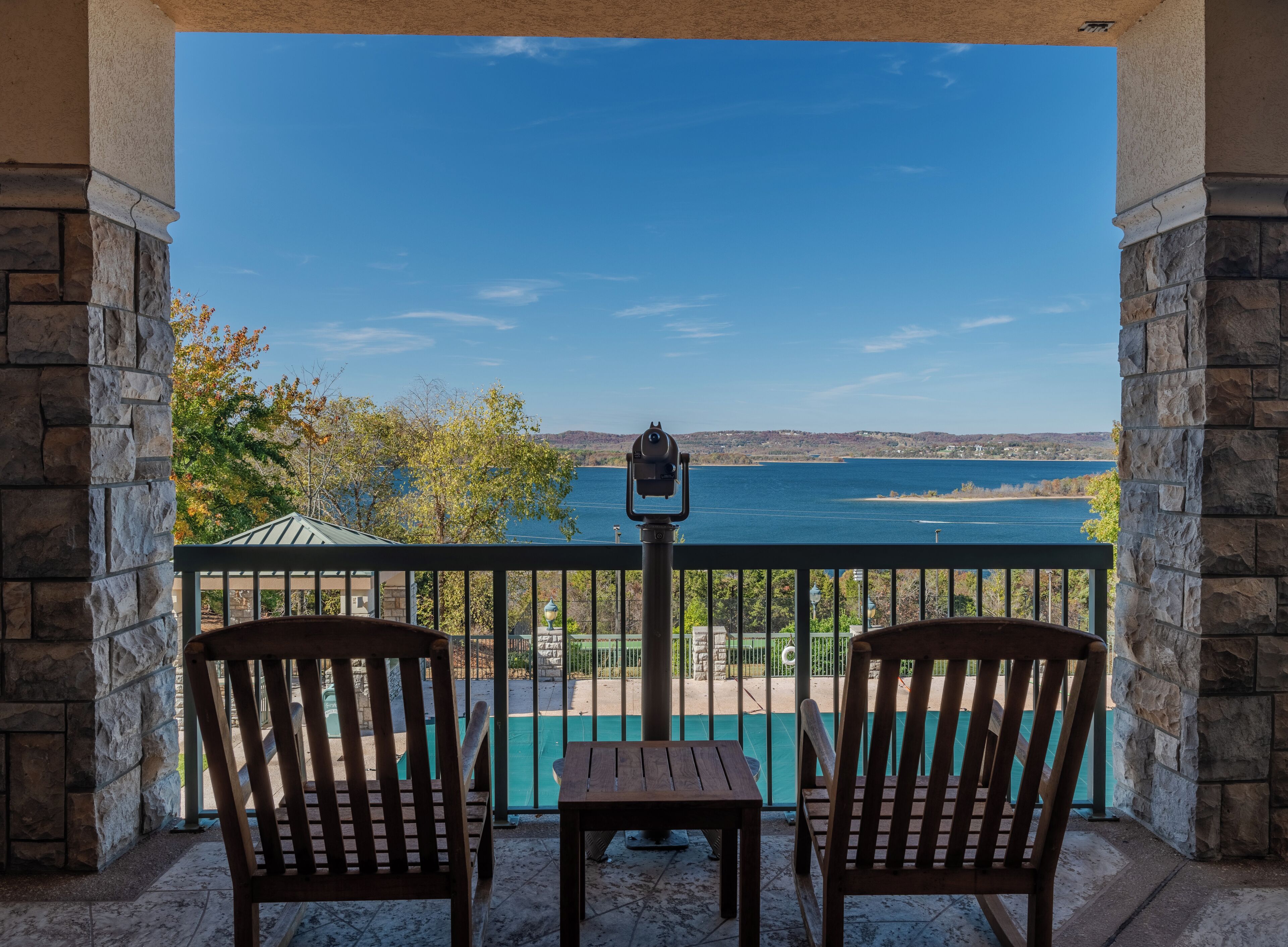 A balcony with two wooden chairs and a small table, overlooking a body of water and distant hills.