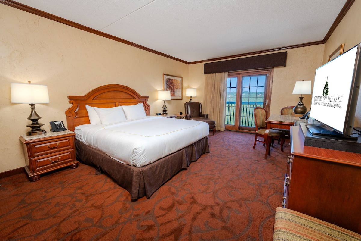 A hotel room with a large bed, two bedside tables with lamps, a desk with a chair, a television, and a view of the outdoors through a window.