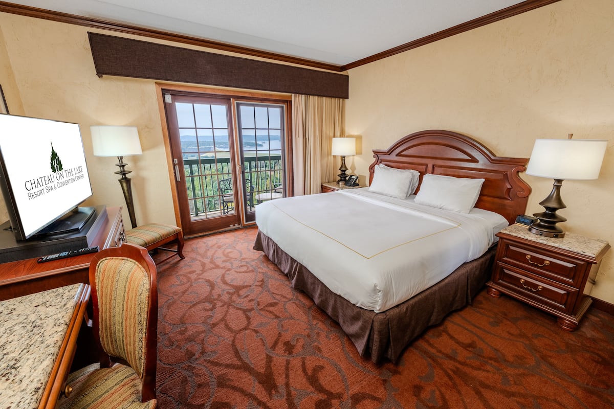 A hotel room with a large bed, a desk with a computer, a chair, and a view of the outdoors through a sliding door.