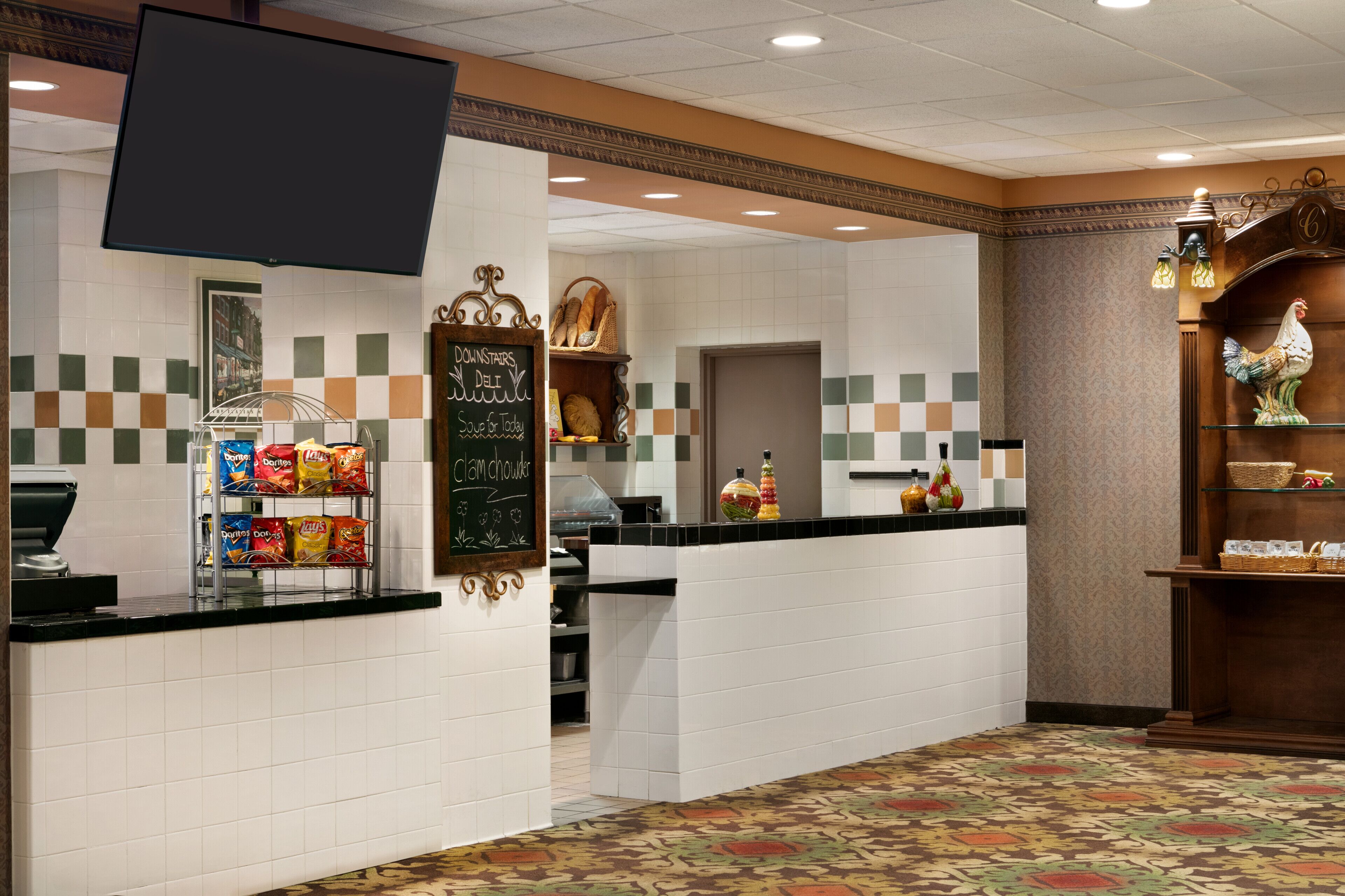 A hotel lobby with a reception desk, a television mounted above it, a chalkboard menu, and a decorative wall with a rooster statue.