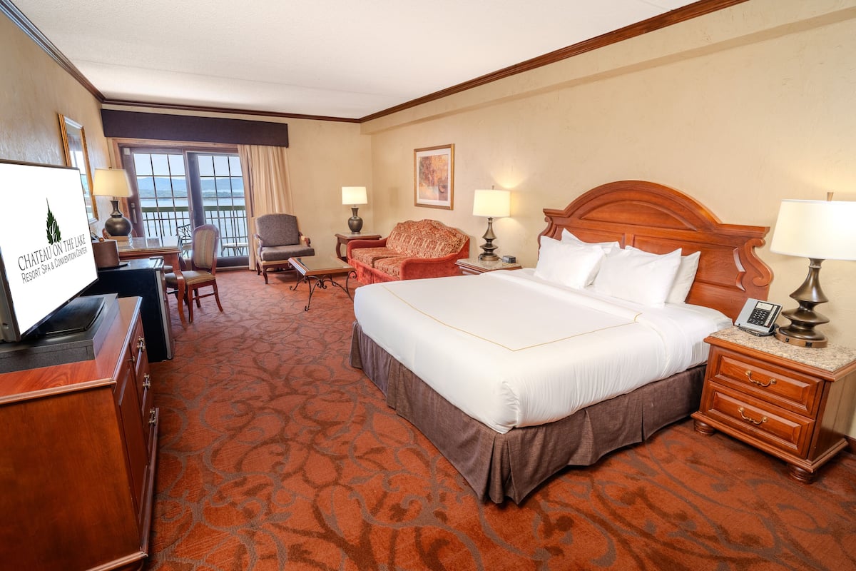 A hotel room with a large bed, a desk with a computer, a chair, a sofa, and a view of the lake.